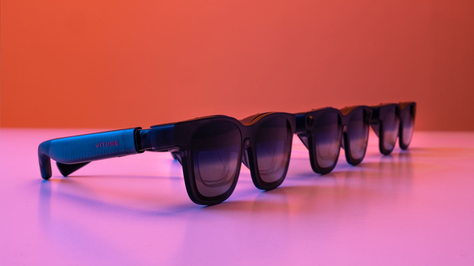 Four pairs of dark VITURE video glasses standing in a row on a pink surface, with the VITURE name on the temple of the nearest pair.