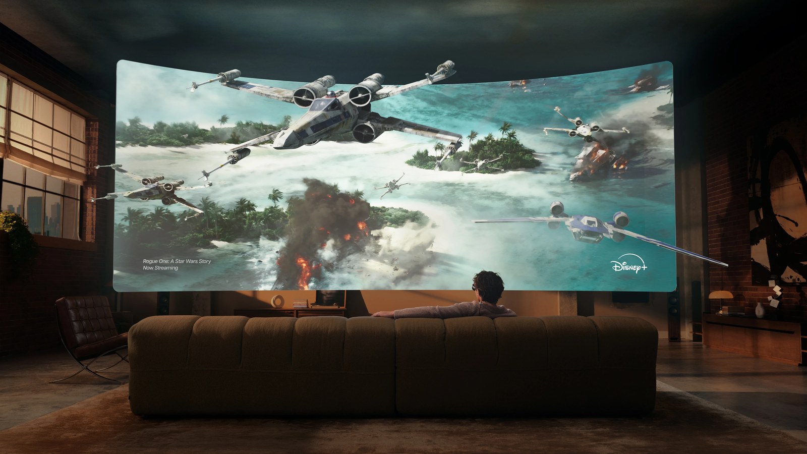 A person on a sofa facing a cinema-sized curved screen that fills the wall of a loft, showing a Star Wars film with the Disney+ logo.