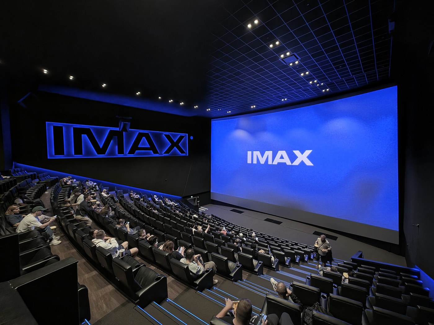 An IMAX auditorium seen from the back rows: a screen that fills the front wall from floor to ceiling, with people in the seats below it.
