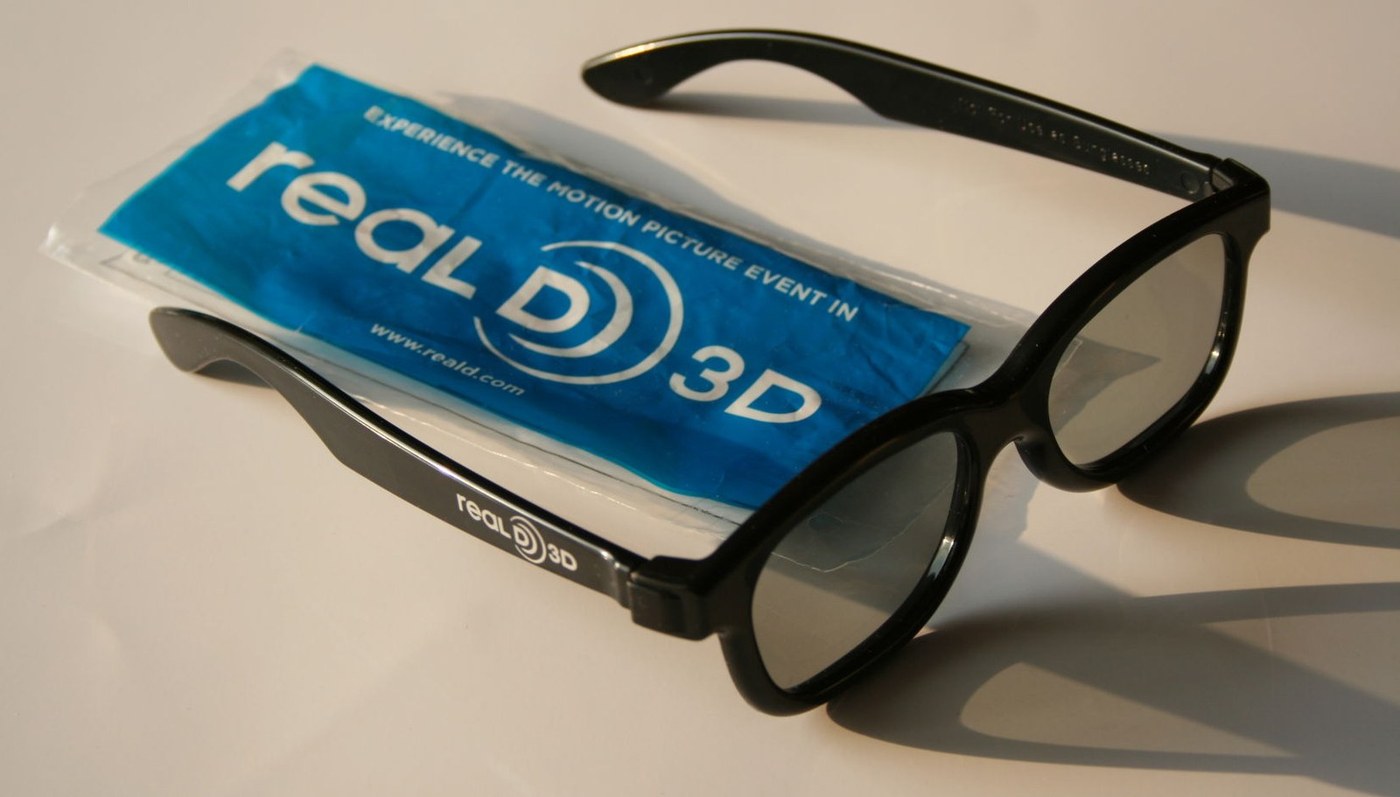 A pair of black RealD 3D glasses lying on the blue plastic bag they came in, both printed with the RealD 3D logo.