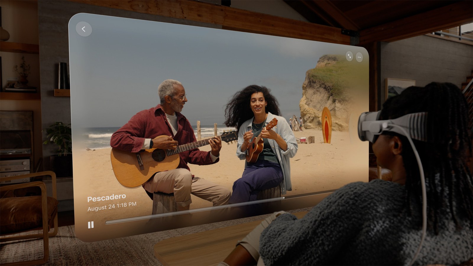 A person wearing an Apple Vision Pro in a living room, looking at a large floating window that shows two people playing guitars on a beach.