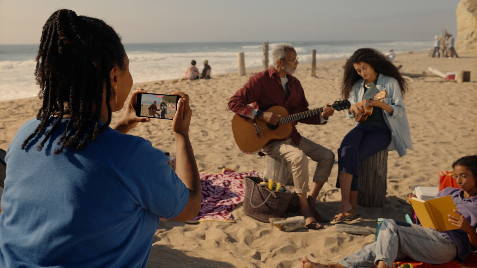 A person on a beach holding an iPhone sideways in both hands, pointing it at two people who are playing guitars a few feet away.
