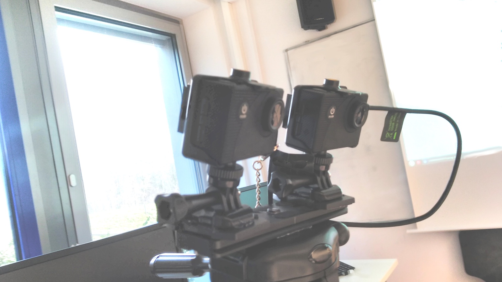 Two small action cameras mounted side by side on one tripod plate, their lenses about an eye's width apart.