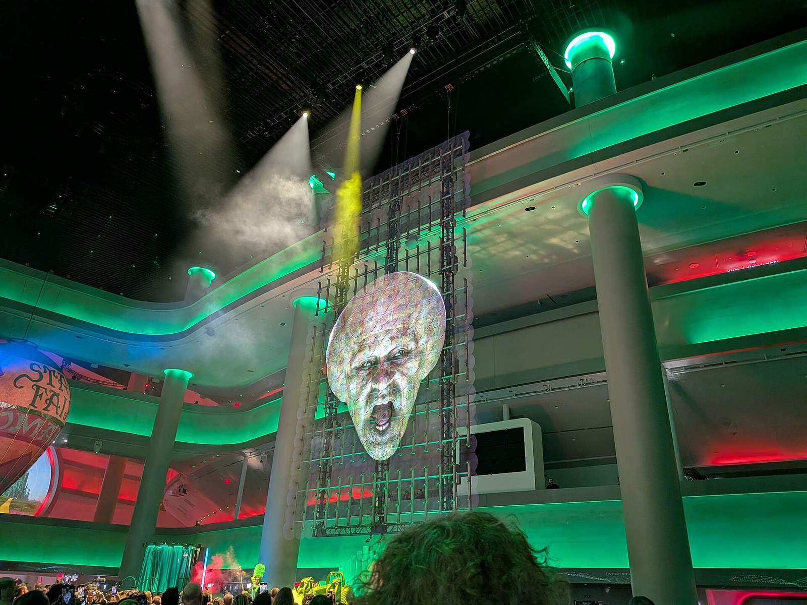 A large face that seems to float in front of a metal frame hung above a crowd in a hall lit green.